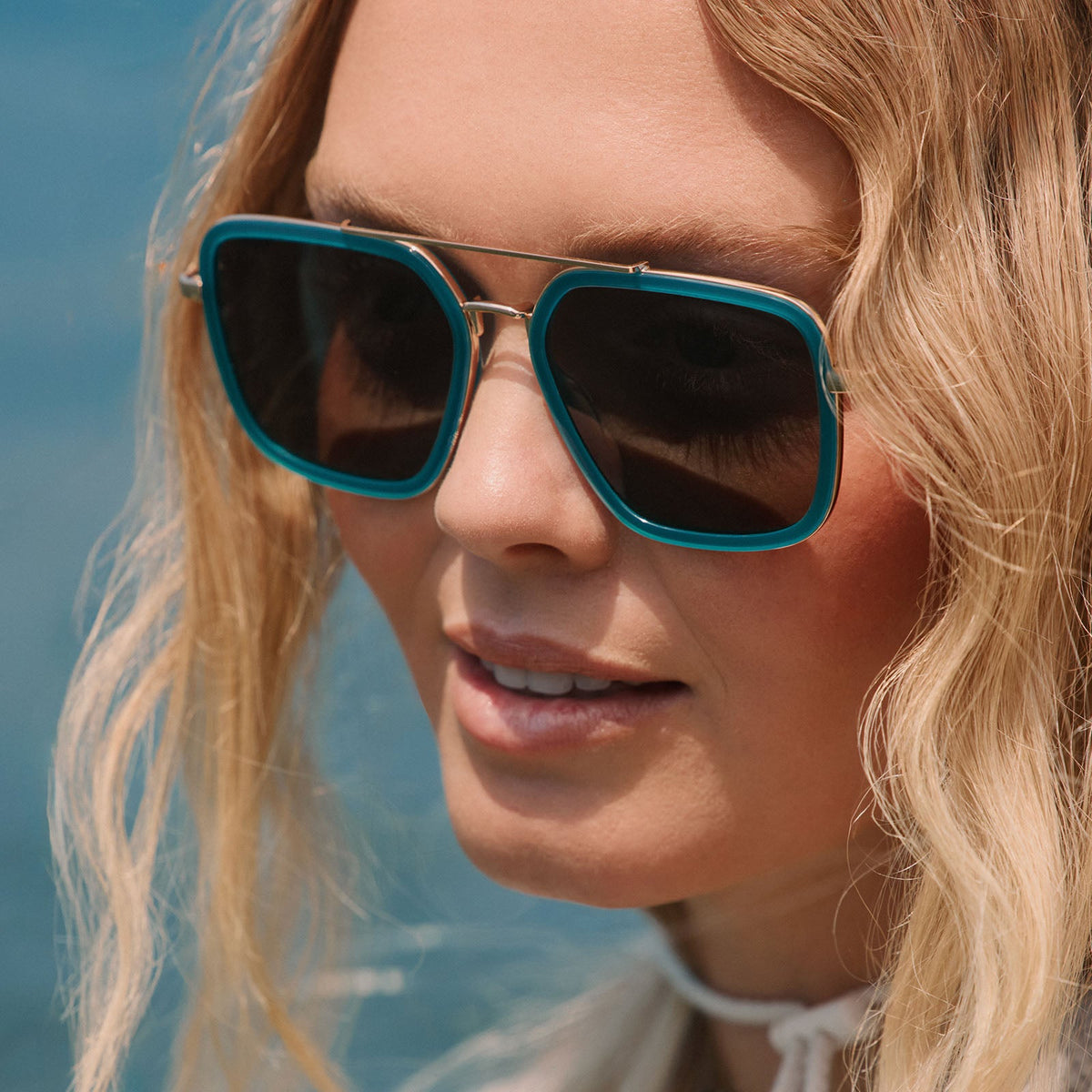lifestyle - blonde female at the beach wearing diff eyewear featuring the jordan navigator sunglasses with a dark teal frame and grey polarized lenses angled view