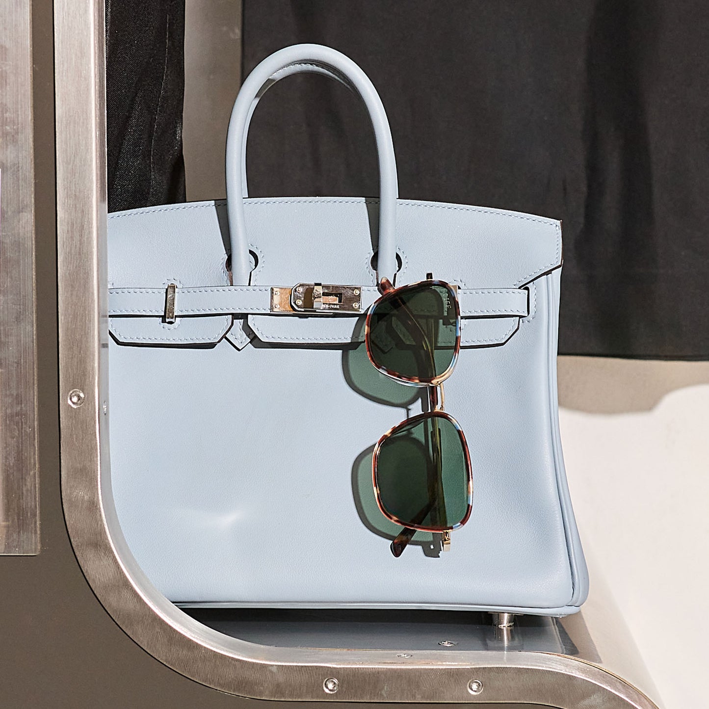 still life of jena sims x diff eyewear hope cat eye sunglasses with a blue color pop frame and g15 polarized lenses hanging on a light blue purse