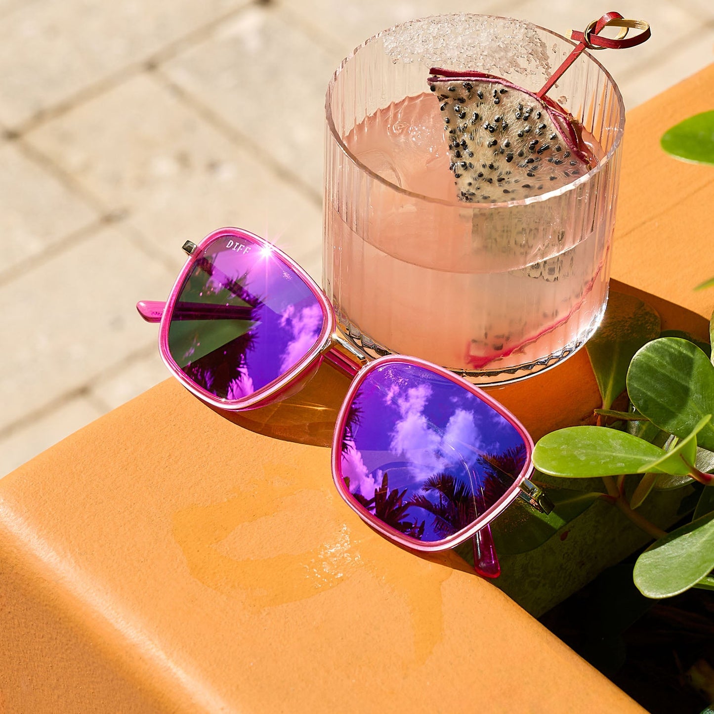 still life - jena sims x diff eyewear the hope cat eye sunglasses with a hot pink and gold frame and hot pink mirror lenses  laying on a table with a beverage