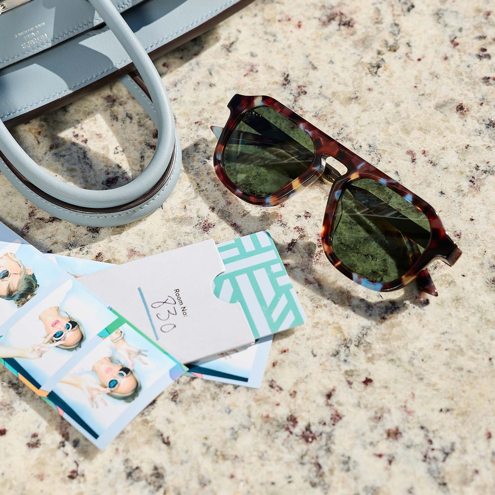 still life - jena sims x diff eyewear the sims navigator sunglasses with a blue color pop with gold metal frame and g15 polarized lenses laying on a table next to a purse