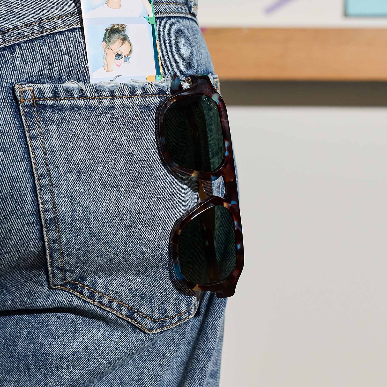 still life - jena sims x diff eyewear the sims navigator sunglasses with a blue color pop with gold metal frame and g15 polarized lenses hanging on her back pocket