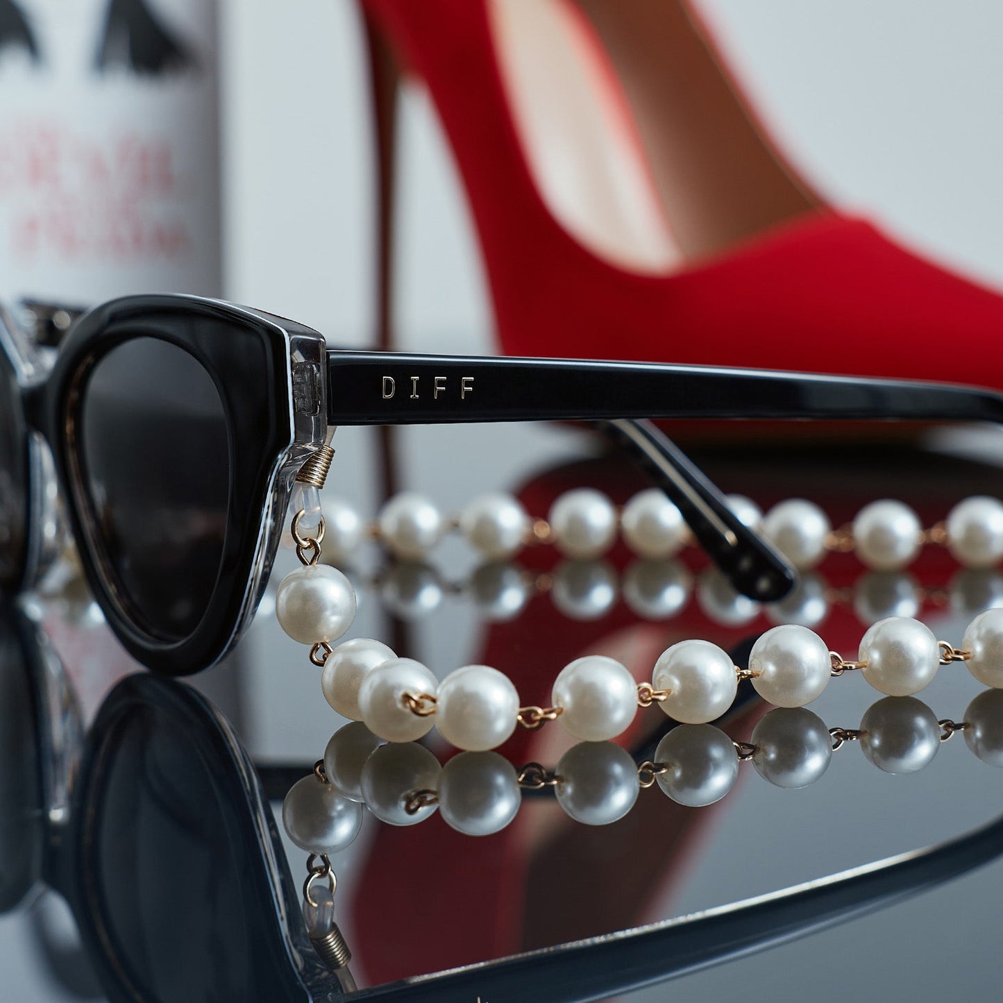 still life - close up view of devil wears prada x diff eyewear andy in black sunglasses with pearl chain with red stiletto shoe in the back ground. 