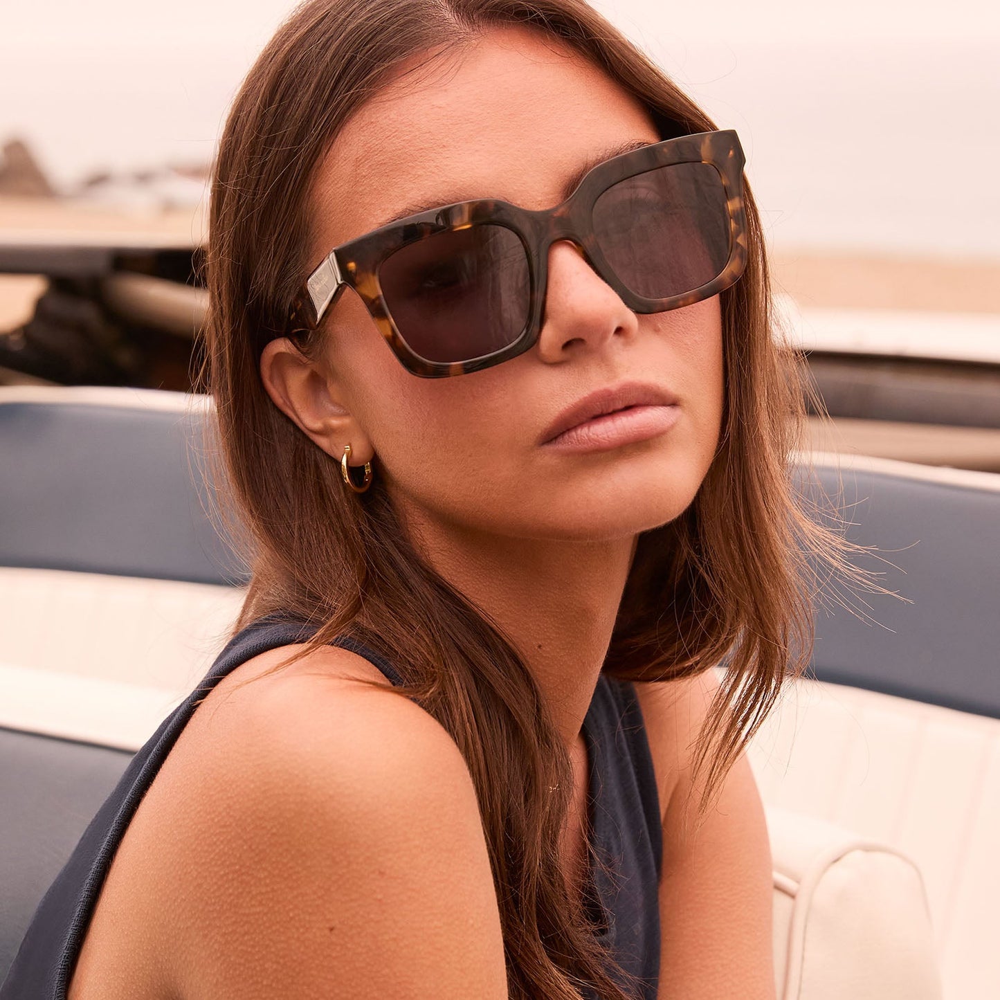 lifestyle - female in her car at the beach wearing travis mathews x diff eyewear the valencia square sunglasses with a tortoise acetate frame and grey polarized lenses angled view