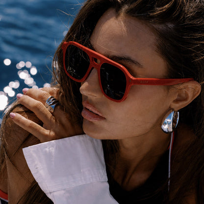 lifestyle - female on a boat wearing beach riot x diff eyewear the coast navigator sunglasses with a red acetate frame and navy blue lenses angled view