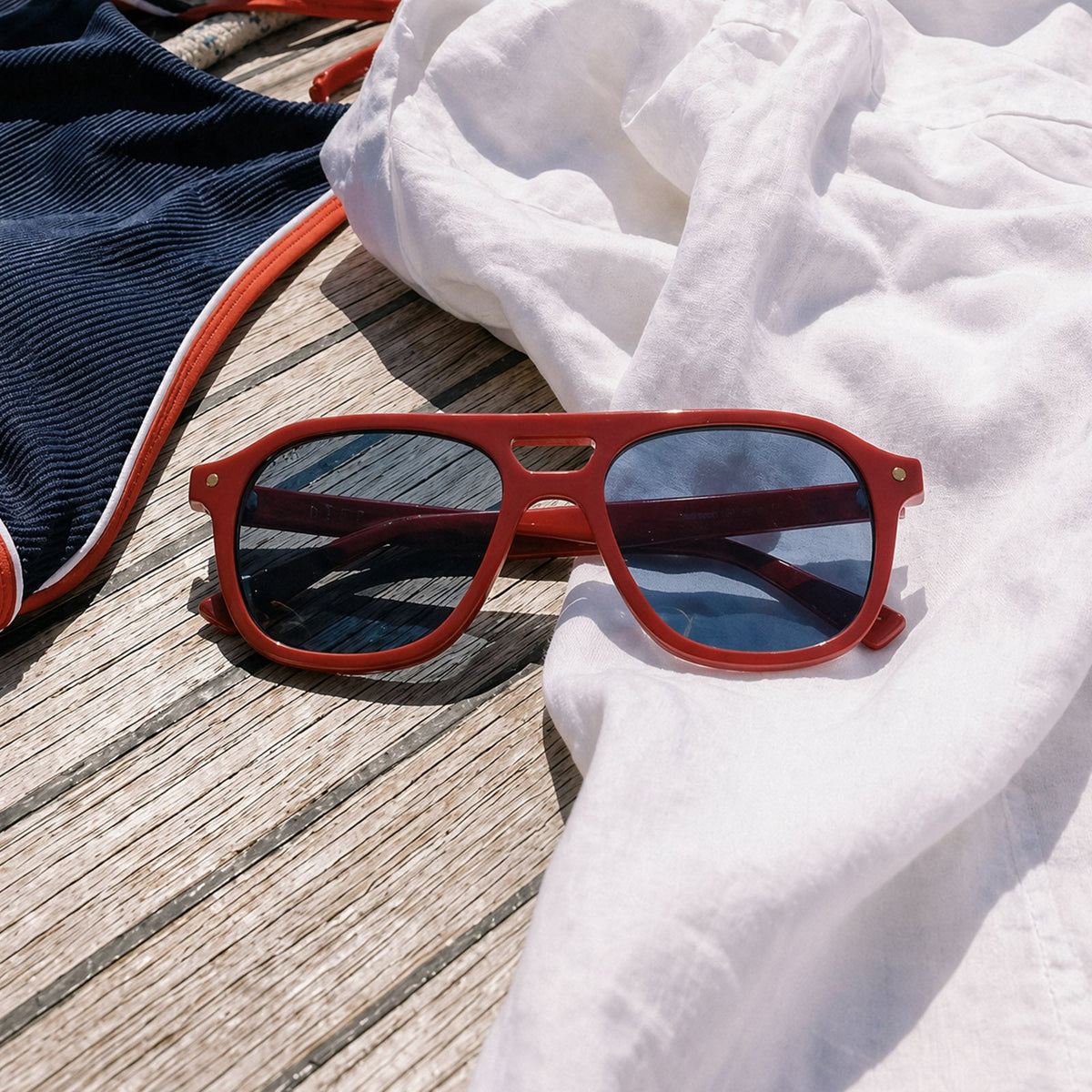still life - beach riot x diff eyewear the coast navigator sunglasses with a red acetate frame and navy blue lenses laying on a deck with clothes
