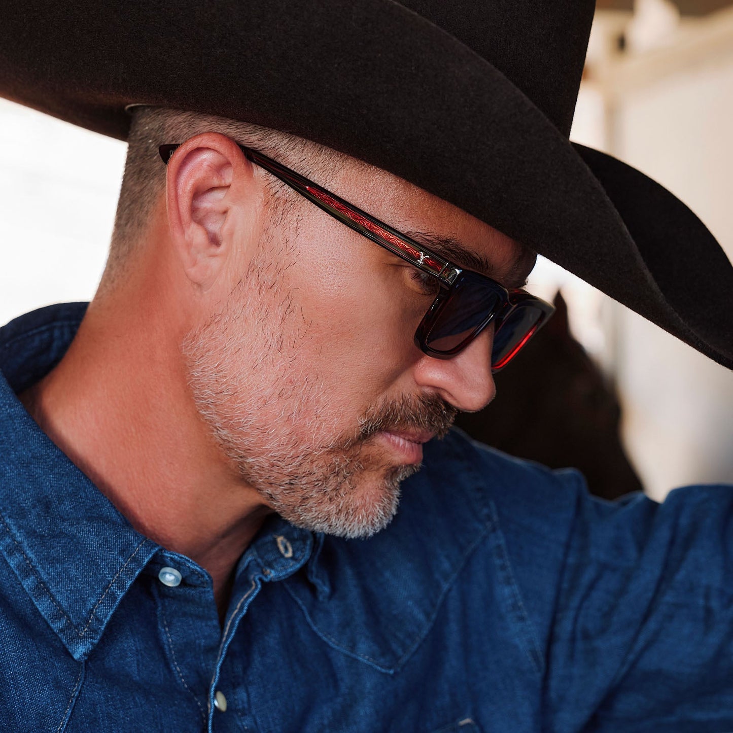 lifestyle - male in horse stables wearing yellowstone x diff eyewear john dutton rectangular sunglasses with a redwood frame and grey polarized lenses side view