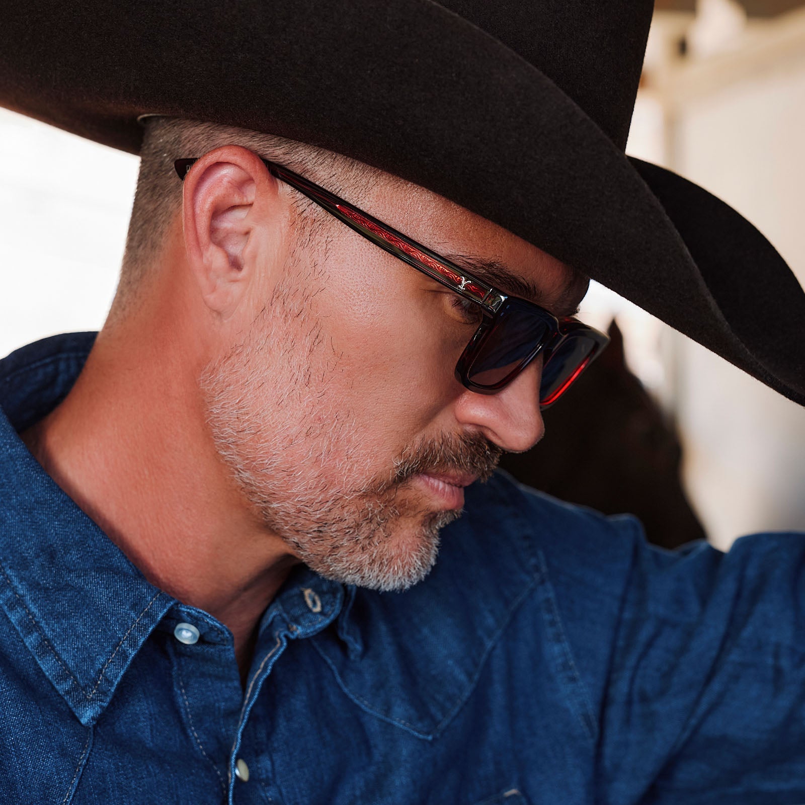 lifestyle - male in horse stables wearing yellowstone x diff eyewear john dutton rectangular sunglasses with a redwood frame and grey polarized lenses side view