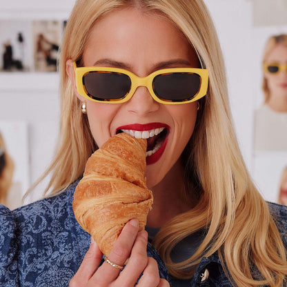 lifestyle - female in photo studio eating a croissant while trying on emily in paris x diff eyewear featuring the julien 2.0 rectangle sunglasses with a citron yellow with tortoise frame and chic grey lenses front view