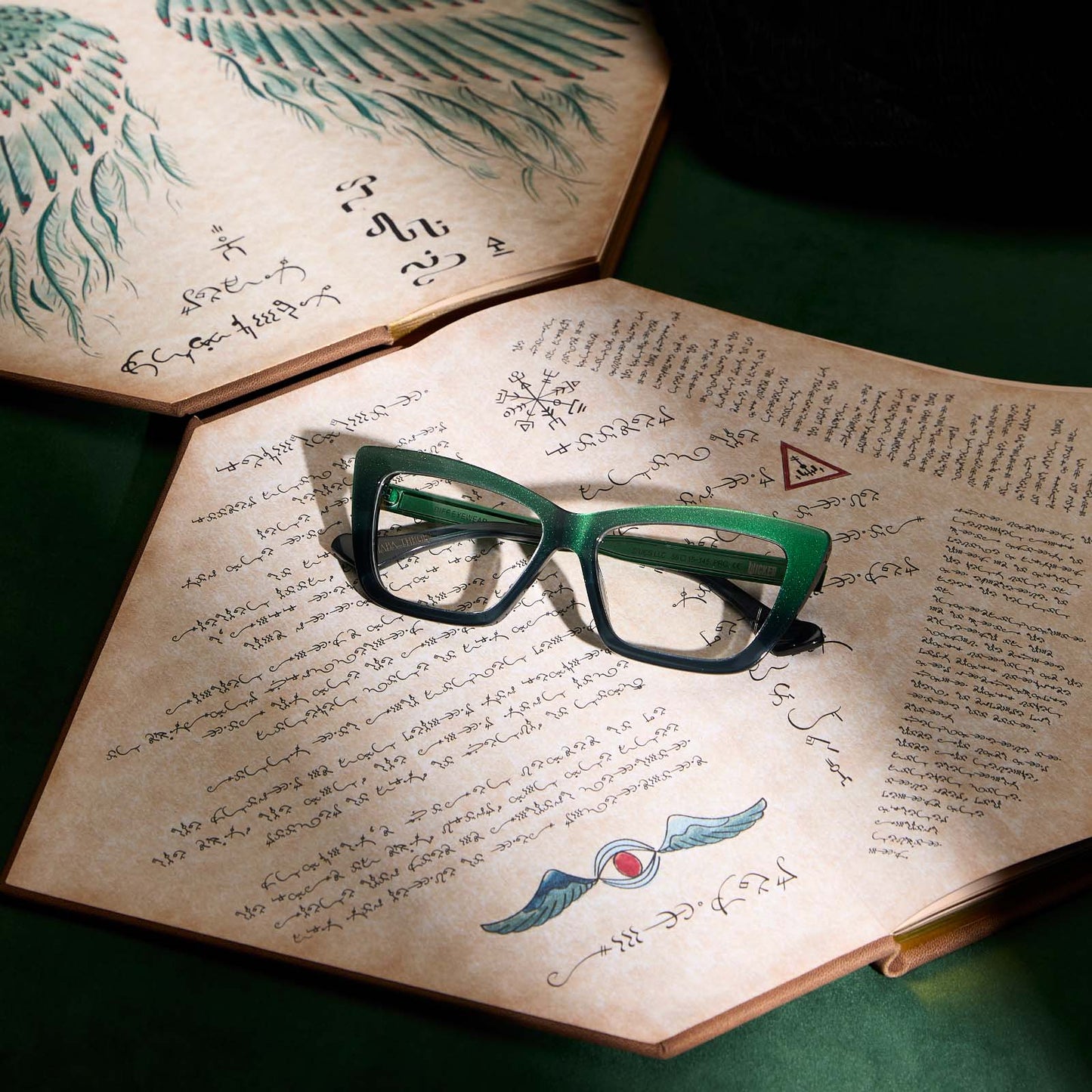 lifestyle - wicked x diff eyewear featuring the elphaba emerald square prescription glasses with a black green gradient frame laying on a manuscript