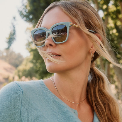 lifestyle - blonde female outdoors wearing diff eyewear bella square oversized sunglasses with a dew drop blue frame, gold temples and brown gradient lenses angled view