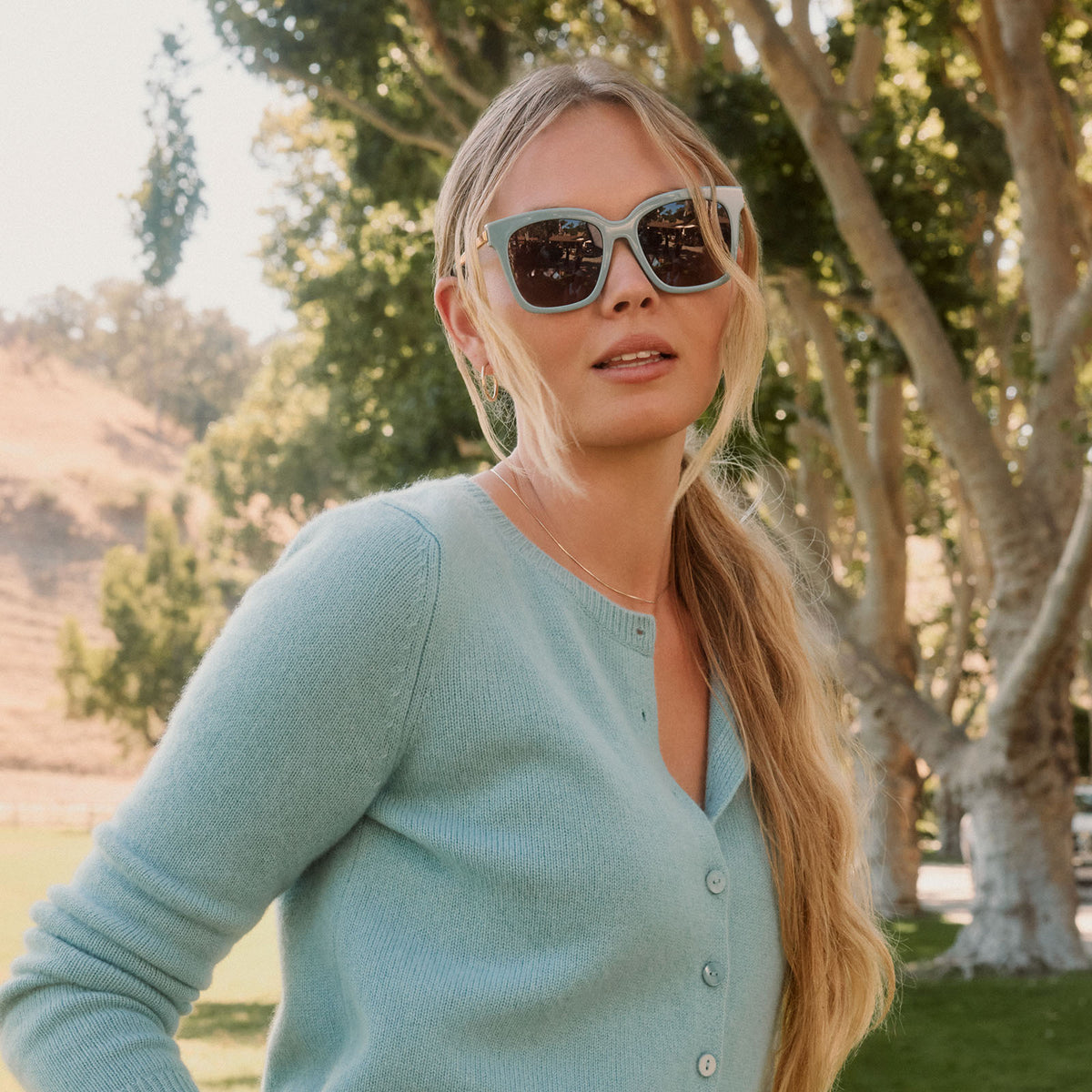 lifestyle - blonde female outdoors wearing diff eyewear bella square oversized sunglasses with a dew drop blue frame, gold temples and brown gradient lenses front view