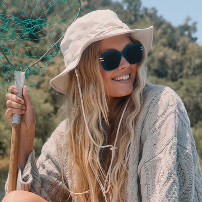 lifestyle - blonde female at the lake wearing diff eyewear dasha square sunglasses with a dark teal acetate frame and grey lenses angled view