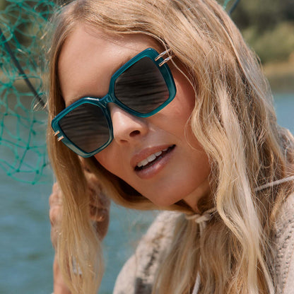 lifestyle - blonde female at the lake wearing diff eyewear dasha square sunglasses with a dark teal acetate frame and grey lenses front view