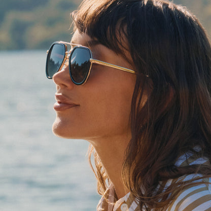 lifestyle - female by a lake wearing diff eyewear featuring the joey round sunglasses with a black & gold metal frame with grey gradient polarized lenses side view