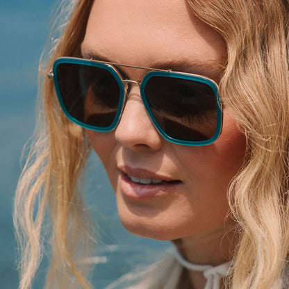 lifestyle - blonde female at the beach wearing diff eyewear featuring the jordan navigator sunglasses with a dark teal frame and grey polarized lenses angled view