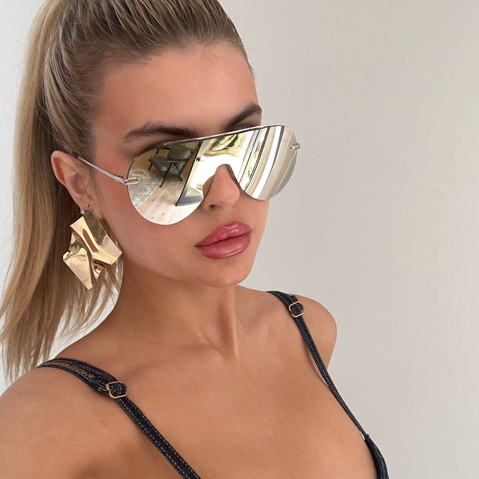 lifestyle - blonde female wearing gold shield sunglasses with matching gold statement earings