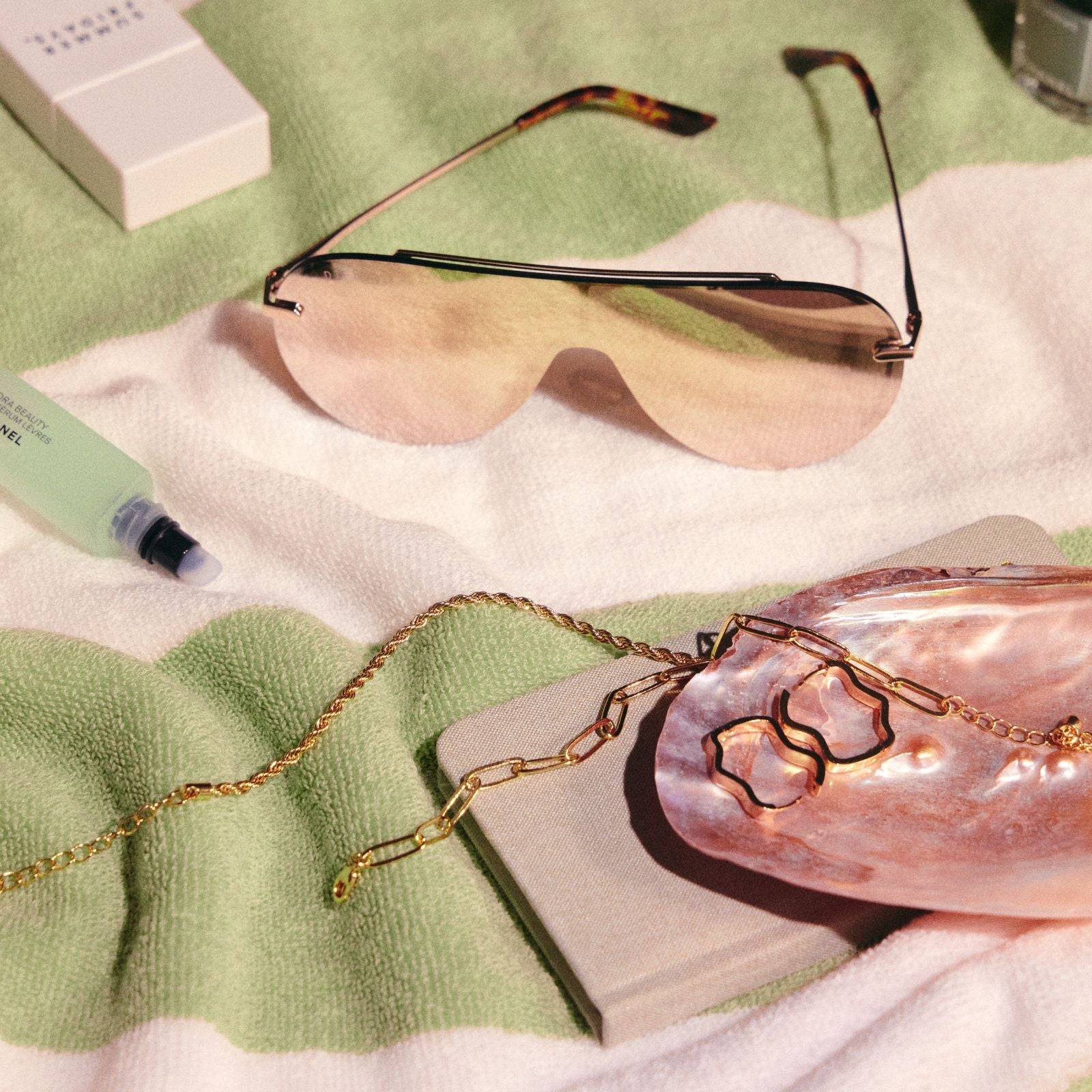 lifestyle - gold shield sunglasses flat lay on green towel with accessories